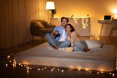 Neşeli bir çift bir yatakta rahatça oturur, film izler ve patlamış mısır paylaşır. Alçak ışık sıcak bir atmosfer yaratıyor, evde rahat bir akşam için mükemmel..