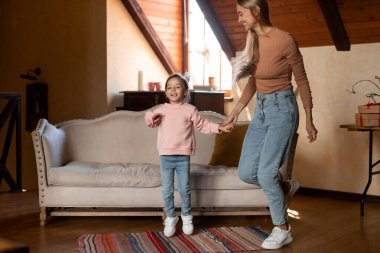 Bir anne ve kızı sıcak ve davetkar bir oturma odasında mutlu bir şekilde zıplıyorlar. Güneş ışığı boşluğu doldurur, neşeli bir atmosfer yaratır..