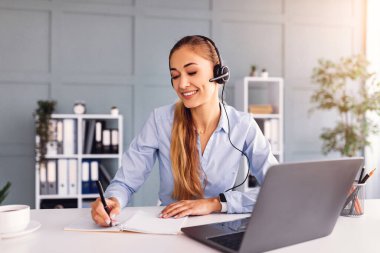 Bir kadın parlak bir ofiste masa başında oturuyor, kulaklık takıyor ve not defterine not alıyor. Masada bir dizüstü bilgisayar ve kahve fincanı da var..