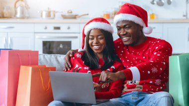 Neşeli bir çift, Noel süveteri ve Noel Baba şapkası giyerek bir koltuğa oturur. İnternetten alışveriş yaparken gülümserler. Etrafları renkli alışveriş torbalarıyla çevrilidir..