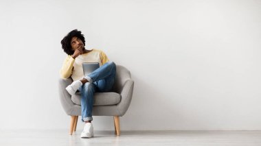Genç siyah bir adam rahat bir sandalyede oturur, ayaklarını koltukta dinlendirirken dijital tabletinde seçenekleri düşünür. Sakin bir ortamda online alışveriş yaptığını düşünürsek, derin düşüncelere dalmış gibi görünüyor..