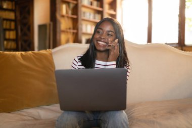 Genç bir kadın kanepede rahatça oturup telefonda konuşurken gülümsüyor. Kitaplarla dolu sıcak ve davetkar bir oturma odasında dizüstü bilgisayarıyla çalışıyor..