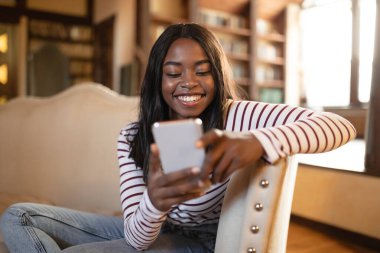 Genç bir kadın rahat bir koltukta oturuyor, akıllı telefonuyla doğal ışıkla dolu sıcak, davetkar bir odada gülümsüyor..