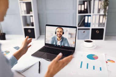 Bir insan sanal bir toplantıya katılır, dizüstü bilgisayardaki bir adamla canlı bir şekilde konuşur. Çalışma alanı, kahve fincanının etrafında not defterleri ve çizelgelerle düzenlenir..