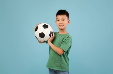 Neşeli genç bir çocuk elinde bir futbol topuyla ayakta duruyor. Yeşil bir gömlek ve kot pantolon giyiyor, spor için coşku ve neşe sergiliyor..