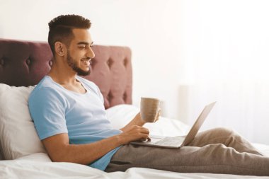 Genç bir adam laptopuna odaklanırken sıcak bir fincan kahveyle yatakta dinlenir. Oda aydınlık ve davetkar. Evde rahat bir çalışma alanı yaratıyor..