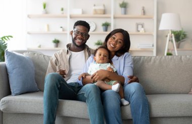 Üç kişilik neşeli bir Afro-Amerikan ailesi koltuklarında oturuyor ve gülümsüyor. Ebeveynler çocuklarını kucaklıyor, evlerinde sıcak ve sevgi dolu bir ortam sergiliyorlar..