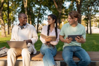 Three friends are seated on a bench in a park, sharing ideas while using laptops and tablets. The sun shines through green trees, creating a lively atmosphere.