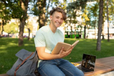 Genç bir adam parlak bir parkta ahşap bir bankta kitap okumaktan hoşlanır. Açık bir günde, dizüstü bilgisayarı ve sırt çantası ağaçların altında dinlenirken yakınlardadır..