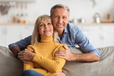 Birbirlerini rahat bir kanepede kucaklarken mutlu bir çift gülümsüyor. Parlak oturma odasında açık renkli ve rahat dekorlu sıcak bir atmosfer var..