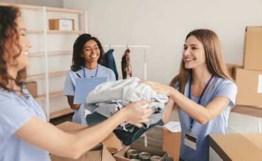 Üç gönüllü aktif olarak bir sosyal merkezde kıyafet sınıflandırıyor ve paylaşıyorlar. Gülümsüyor ve işlerine dalmış durumdalar. Olumlu bir takım çalışması ve destek ortamı yaratıyorlar..