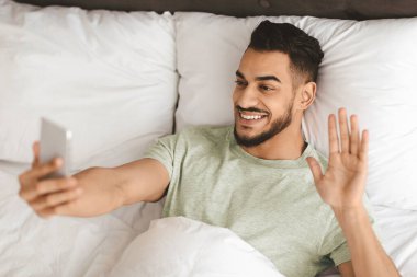 A man with a beard is smiling and waving while holding a smartphone in bed. He is wearing a light green shirt and appears to be relaxed, enjoying a conversation.