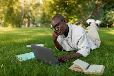 A man lies on the grass in a sunny park, focusing on his laptop while surrounded by open books. He appears relaxed and engaged in his work amidst greenery.