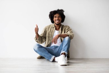 Genç bir Afro-Amerikan adam yerde oturuyor, gülümsüyor ve baş parmak işareti yapıyor. Rahat, günlük kıyafetler giyiyor ve beyaz bir duvarın yanında bir anlık pozitifliğin tadını çıkarıyor..
