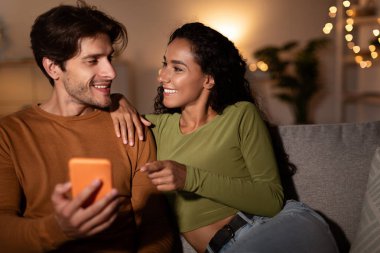 Akşamları, bir çift evdeki kanepede oturur, akıllı telefona bakar. Kadın, kocasıyla sıcak ve samimi bir sahne yaratırken ekrandaki bir şeye işaret ediyor..