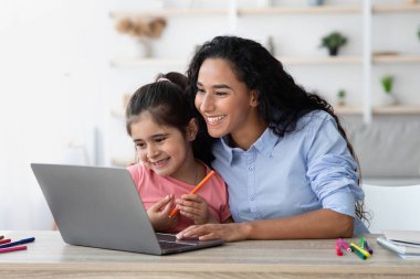 Bir anne ve küçük kızı bir masada oturuyor, gülümsüyor ve dizüstü bilgisayarla meşgul. Konforlu ev dekorunun etrafa dağılmış renkli sanat malzemeleri eğlenceli bir öğrenme aktivitesi olduğunu gösteriyor..