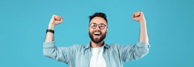 Gözlüklü ve sakallı bir adam heyecanla kollarını kaldırıyor. Yüzünde büyük bir gülümseme var, coşku gösteriyor. Arka plan masmavi, neşeli bir hava katıyor..