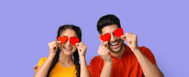 Genç bir çift, kırmızı kalp şekillerini gözlerinin önünde tutarak neşe içinde poz veriyor. Neşeli ifadeleri ve renkli kıyafetleri, neşeli ve neşeli bir anı vurguluyor..