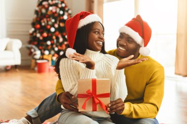 Bir çift yerde oturup gülümsüyor ve birlikte vakit geçiriyorlar. Noel Baba şapkası takıyorlar. Arka planda süslemeli bir Noel ağacı var. Bir hediye onların ellerinde..