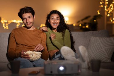 Couple relaxing on the sofa at home watching a movie. The wife is enjoying the film and smiling, while the husband appears bored with the choice. Its a cozy family weekend.