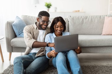 Afrika kökenli Amerikalı bir çift oturma odalarında halının üzerinde oturuyor. Bir dizüstü bilgisayar kullanıyorlar, gülümsüyorlar ve çevrimiçi içerikten keyif alırken ekranı gösteriyorlar..
