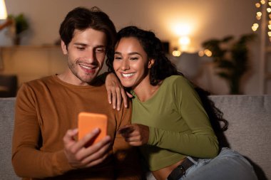 Çift evdeki kanepelerinde oturup cep telefonlarındaki yeni bir başvuruyu zevkle inceliyor. Kadın heyecan verici bir şekilde ekranı işaret ediyor, eşiyle harika uygulama önerilerini paylaşıyor..