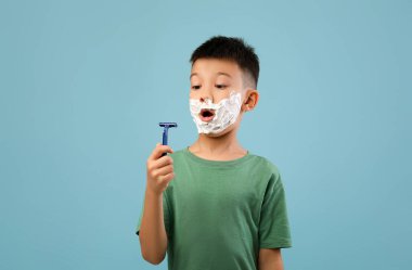 Yüzünde tıraş kremi olan genç bir çocuk elinde jilet tutarken şaşırmış görünüyor. Yeşil bir gömlek giyiyor ve yumuşak mavi bir arka plana karşı tuhaf bir ifadesi var..