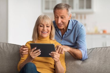 Bir erkek ve bir kadın birlikte bir tablete bakarken keyifli bir anı paylaşıyorlar. Parlak bir oturma odasındaki koltukta oturup aralarındaki mutluluğu ve bağlantıyı gösteriyorlar..