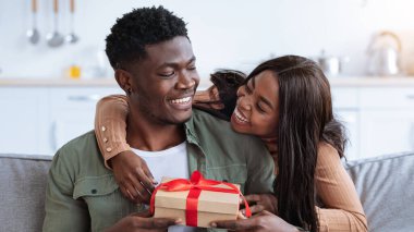 A young couple sits on a couch in their cozy living room, smiling and laughing as they exchange gifts.