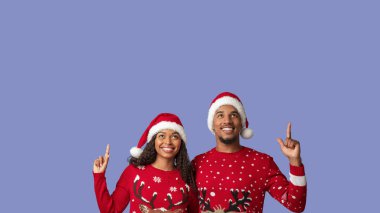 Neşeli bir çift yukarı bakarken kırmızı Noel süveteri ve Noel Baba şapkası giyer. Parlak bir şekilde gülümserler, bayram sezonu için heyecanı paylaşırlar. Arka plan katı bir açık renk..