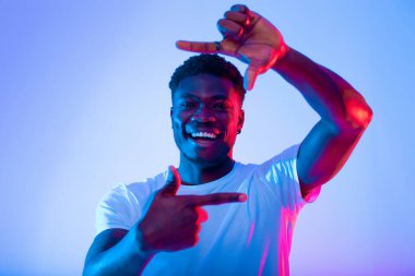 Neşeli bir adam elleri ile poz veriyor, büyük bir gülümseme sergiliyor. Canlı mavi ve pembe ışık arka planda canlı bir atmosfer yaratıyor..