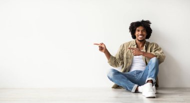 Genç Afro-Amerikalı adam yere oturup kameraya bakıyor, gülümsüyor ve işaret ediyor. O, düz beyaz bir duvarla birlikte parlak bir kapalı alanda. Reklam için bir sürü yer var..