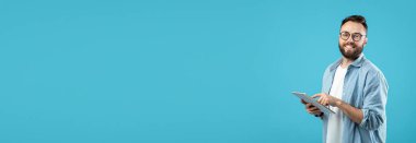 Sakallı ve gözlüklü genç bir adam elinde bir tablet tutarken gülümsüyor. Canlı bir mavi duvara karşı duruyor, mutlu görünüyor ve cihazla meşgul..