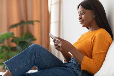 Genç bir kadın oturma odasındaki kanepede dinleniyor, akıllı telefonuyla meşgul. Kablosuz kulaklık takıyor ve iç mekan bitkileri ve sıcak aydınlatmalarla dolu huzurlu bir anın tadını çıkarıyor..