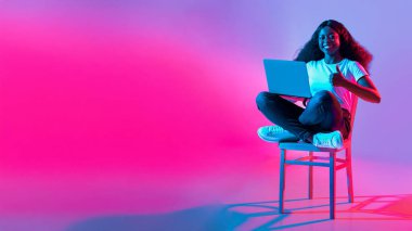Genç bir kadın dizüstü bilgisayar kullanarak bir sandalyeye bağlanmış oturuyor ve gülümsüyor. Arkaplanda canlı pembe ve mavi renkler var. Üretkenlik için mükemmel bir atmosfer yaratıyorlar..
