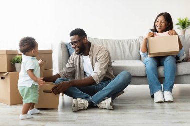 Neşeli Afro-Amerikan ailesi yeni bir daireye taşınıp, boks malzemelerini çıkararak ve birlikte vakit geçirerek eğleniyor. Küçük çocuk babasına yardım ederken annesi kanepede gülümsüyor..