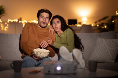 A couple sits on a couch in their living room, watching a horror movie with a projector. They look shocked and frightened while enjoying popcorn in a cozy, dimly lit space.