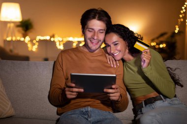 Mutlu çift akşamları kanepelerinde oturup internetten alışveriş yapmak için bir tablet ve kredi kartı kullanıyor. Dijital bir mağazayı keşfederken gülücüklerini paylaşıyorlar, birlikte vakit geçiriyorlar..