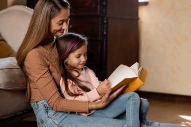 A mother and her young daughter sit comfortably on the floor of a warm living room, sharing a book. The child points to the pages as the mother smiles and reads aloud, creating a bonding moment.