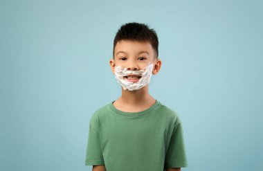 Yeşil gömlek giyen bir çocuk açık mavi bir arka plana karşı duruyor. Yüzünde tıraş köpüğü var. Gülümserken neşeli ve esprili bir ifade oluşturuyor..
