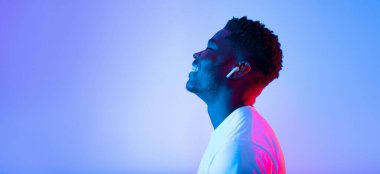 Genç bir adam kablosuz kulaklıklardan müzik dinlerken gözleri kapalı gülümser. Arka planda canlı mavi ve pembe renkler var, rahat bir atmosfer yaratıyor..