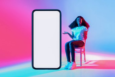 Kıvırcık saçlı genç bir kadın bir sandalyede oturuyor ve boş bir akıllı telefon ekranına doğru hareket ederken gülümsüyor. Arkaplanda canlı mavi ve pembe ışıklar var..