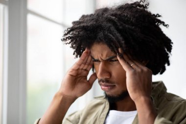 Genç bir siyah adam büyük bir pencerenin yanında duruyor, gözle görülür şekilde üzgün ve şakaklarını tutuyor. Baş ağrısından muzdarip gibi görünüyor, evde rahatsızlık belirtileri gösteriyor..