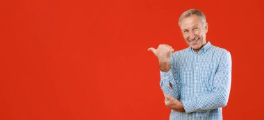 Gri saçlı bir adam kırmızı arka planda gülümsüyor. Sağ eliyle yanındaki bölgeyi işaret ediyor. Günlük kıyafetleri olay yerinin dostane atmosferine katkı sağlıyor..