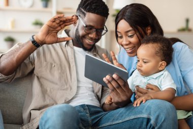 Gülümseyen siyahi ebeveynler video görüşmesi için tablet kullanırken bebeklerini tutarlar. Ekrana el sallarlar, aile üyeleriyle oturma odalarının konforundan iletişim kurarlar..