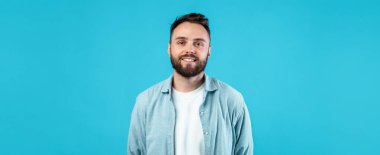 Genç bir adam, canlı mavi bir arkaplan önünde güler yüzlü bir şekilde duruyor. Beyaz bir tişörtün üzerine gündelik, açık renk bir gömlek giyer, rahat bir stili sergiler..