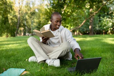 Gülümseyen genç bir adam parktaki yeşil çimenlerde oturuyor, bir elinde kitap, diğerinde dizüstü bilgisayar kullanıyor. Doğanın ortasında güneşlenmekten hoşlanıyor..
