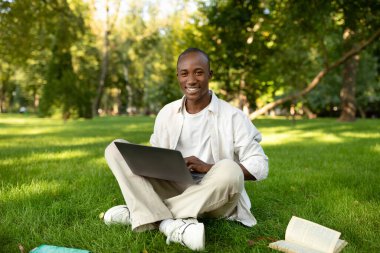 A young man sits cross-legged on the grass in a park, smiling as he types on his laptop. A book lies open nearby, and sunlight filters through the trees.