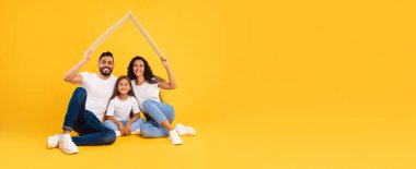 Üç kişilik mutlu bir aile, ellerinde karton bir çatıyla yerde oturuyor. Eşleşen beyaz gömlek ve kot pantolon giyiyorlar. Gülüşleri neşe ve beraberliği gösteriyor..