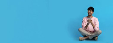 Neşeli genç bir adam mavi bir arka planda bağdaş kurmuş, elinde iki eliyle bir bardak tutuyor. Günlük kıyafetler giyiyor ve rahatlamış görünüyor, zamanının tadını çıkarıyor..